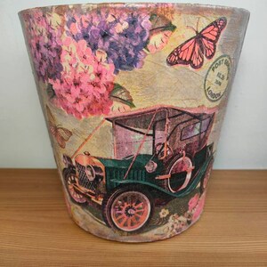 May include: A vintage-style planter with a floral design featuring pink and purple flowers, a butterfly, and a vintage car. The planter has a textured surface and a brown, white, and pink colour scheme. The planter has a stamp that says "Post Office" and "London" on it.