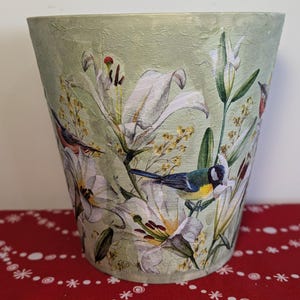 May include: A green ceramic planter with a floral design featuring white lilies, green leaves, and colourful birds. The planter has a textured surface.