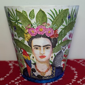 May include: A white decorative planter with a portrait of Frida Kahlo, surrounded by tropical foliage, a parrot, and a lemur. The planter features a floral crown and a necklace. The design is colourful and detailed, with a focus on the iconic artist.