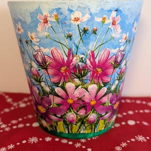 May include: A decorative flower pot with a blue sky background and a vibrant floral design. The pot features large pink and purple cosmos flowers, with white flowers and green stems. The rim is painted light blue, and the base has a green trim.