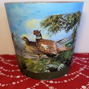 May include: A ceramic planter with a blue sky and green tree background. The planter features a colourful illustration of three pheasants in a woodland setting.