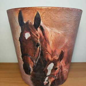 May include: A decorative, tapered vase featuring a detailed image of a brown horse and foal. The horse and foal have white markings on their faces and are wearing bridles. The vase has a textured, metallic finish.