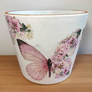 May include: White ceramic planter with a pink butterfly and floral design. The planter has a gold rim.
