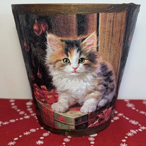 May include: A ceramic planter with a festive Christmas design featuring a cute kitten sitting on a stack of presents. The planter has a brown and gold rim and is decorated with a Christmas tree, baubles, and a red ribbon.
