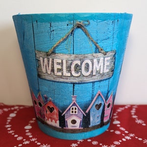 May include: A turquoise blue planter with a distressed woodgrain finish. The planter features a wooden sign that says "WELCOME" hanging from a twine rope. The planter is decorated with a row of colourful birdhouses.