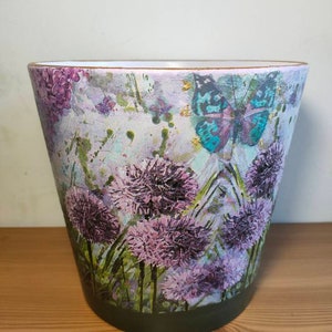 May include: A ceramic planter with a floral design featuring purple flowers, green leaves, and a blue butterfly. The planter has a dark green rim.