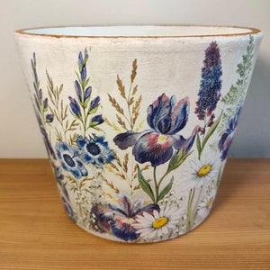 May include: White ceramic flower pot with a floral design featuring blue, purple, and white flowers. The pot has a slightly distressed look and is perfect for adding a touch of nature to any home.