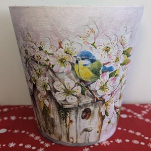 May include: A white ceramic planter featuring a painted design of a bluebird perched on a birdhouse surrounded by white blossoms.