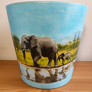 May include: A blue ceramic planter featuring a painted image of elephants in a savanna setting. The planter has a gold rim and a reflective surface.