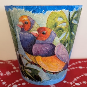 May include: A decorative flower pot with a bird design. The birds have orange heads, purple bodies, and yellow bellies. The pot has a blue rim and a light blue background with green leaves.