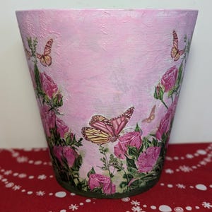 May include: A pink ceramic planter with a floral design featuring roses and butterflies. The planter has a textured surface and a slightly distressed look.