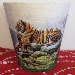 May include: A decorative flower pot with a detailed image of a tiger and cub on a rocky landscape. The pot has a white textured background and a green rim. The tiger has orange and black stripes.