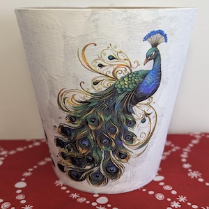 May include: A tapered, off-white planter featuring a detailed, colourful illustration of a peacock. The bird displays vibrant blue, green, and gold plumage with intricate feather patterns. The planter has a textured, matte finish on its exterior surface.
