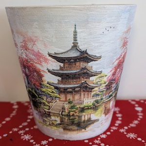 May include: A white ceramic planter featuring a painted image of a Japanese pagoda surrounded by cherry blossom trees and a pond. The pagoda is brown with a green roof and is reflected in the water.