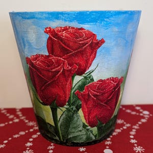 May include: A blue painted flower pot with a design of three red roses with green leaves. The pot is sitting on a red surface with a white pattern.