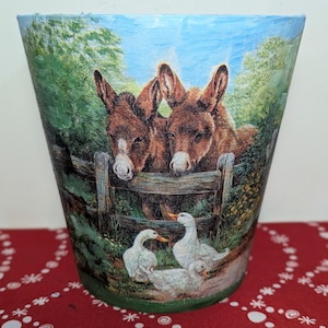 May include: A ceramic planter featuring a painted scene of two brown donkeys peering over a wooden fence with two white ducks in the foreground. The planter has a blue sky and green foliage background.