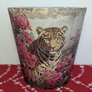 May include: A decorative waste bin with a leopard design surrounded by pink flowers. The bin has a tapered shape and a textured, metallic finish. The leopard's face is detailed with spots and stripes, and the flowers are a vibrant pink.