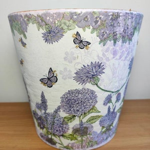 May include: A white ceramic planter with a floral design featuring purple flowers and butterflies. The planter has a textured surface and a slightly flared rim.