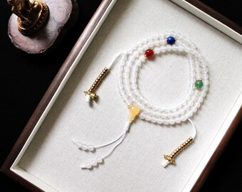 May include: White prayer beads with gold accents and four colored beads. The beads are arranged in a circle and are strung on a white cord. The beads are in a wooden box with a white fabric lining.