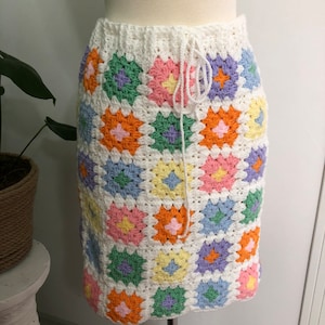 Op de afbeelding: Een witte gehaakte rok met een kleurrijk granny square patroon. De rok heeft een trekkoord in de taille en is gemaakt met verschillende pastelkleuren, waaronder roze, blauw, groen en geel.