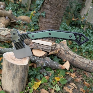 May include: A tactical hatchet featuring a matte grey metal blade, a carbon fibre patterned handle frame, and a textured olive green grip insert secured with three black screws. The tool includes a triangular cutout at the handle end.