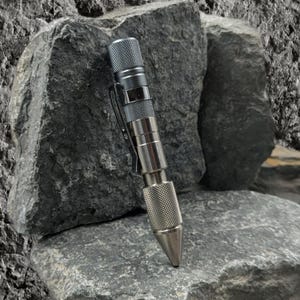 May include: A silver metal pen with a textured grip and a pocket clip. The pen has a conical tip and is displayed against a backdrop of gray rocks. The pen's design suggests a focus on durability and precision.