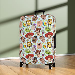 May include: A light blue suitcase cover with a repeating pattern of cartoon character faces, including a cowboy, a space ranger, and a pig. The design includes stars, a rocket, and a yellow ball. The suitcase has a black handle and wheels.