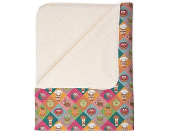 May include: A folded sherpa fleece blanket featuring a colorful diamond pattern filled with various Muppet character faces including Kermit, Fozzie, Animal, and the Swedish Chef, set against a cream-colored textured interior lining.