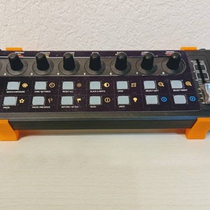 May include: A black Behringer audio interface with orange supports. It features multiple knobs, buttons, and a slider. The interface has labels such as "MIC MODE" and "LAYER".