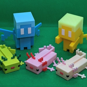 May include: A collection of colorful, 3D-printed Minecraft figures. The set includes two winged creatures in blue and yellow, and three axolotl figures in green, pink, and off-white. The figures are displayed on a green surface.