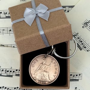 May include: A vintage 1964 British penny coin keychain presented in a small brown gift box with a silver ribbon and bow. The coin features a seated Britannia design and is attached to a silver keyring.