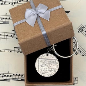 May include: A silver keychain featuring a coin with the words "TEN PENCE" and a heraldic design, presented in a brown gift box tied with a silver ribbon. The box is open, revealing the coin and a black interior.