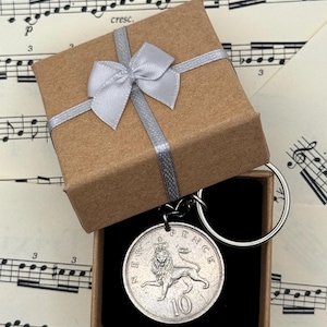 May include: A silver coin keychain with a lion emblem, presented in a small brown gift box with a silver ribbon and bow. The keychain is displayed against a backdrop of sheet music.