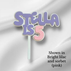 May include: A cake topper with the text "STELLA IS 5" in a bubble font. The letters "STELLA" are lilac, the number "5" is pink, and the text is outlined in white. The topper is on a white stick.