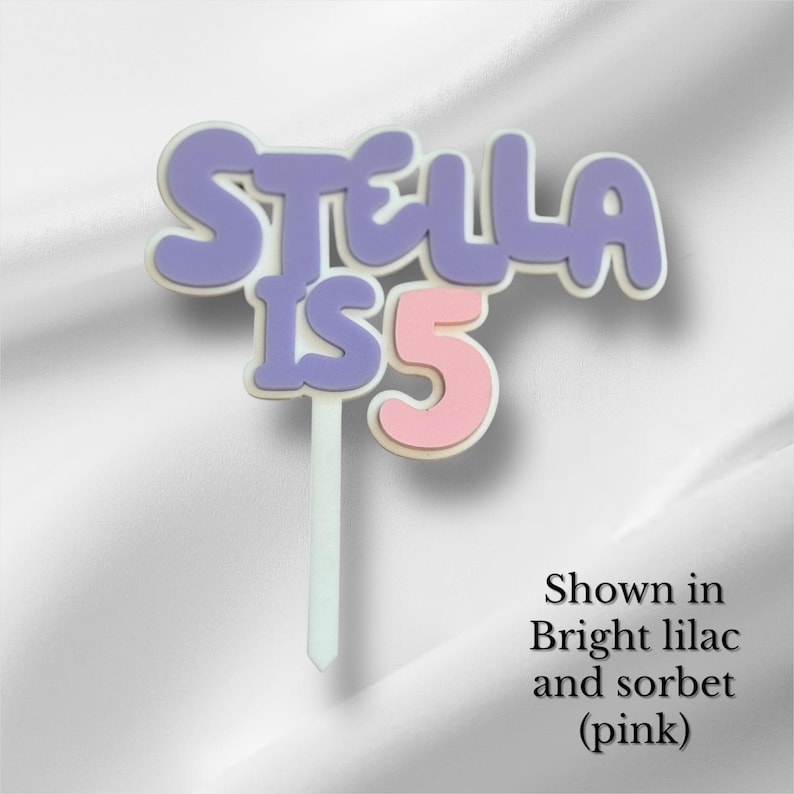 May include: A cake topper with the text "STELLA IS 5" in a bubble font. The letters "STELLA" are lilac, the number "5" is pink, and the text is outlined in white. The topper is on a white stick.