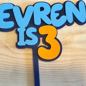 May include: A blue and orange cake topper with the text "EVREN IS 3". The letters are in a bubble font with a dark blue outline. The number 3 is orange. The topper is on a wooden surface.
