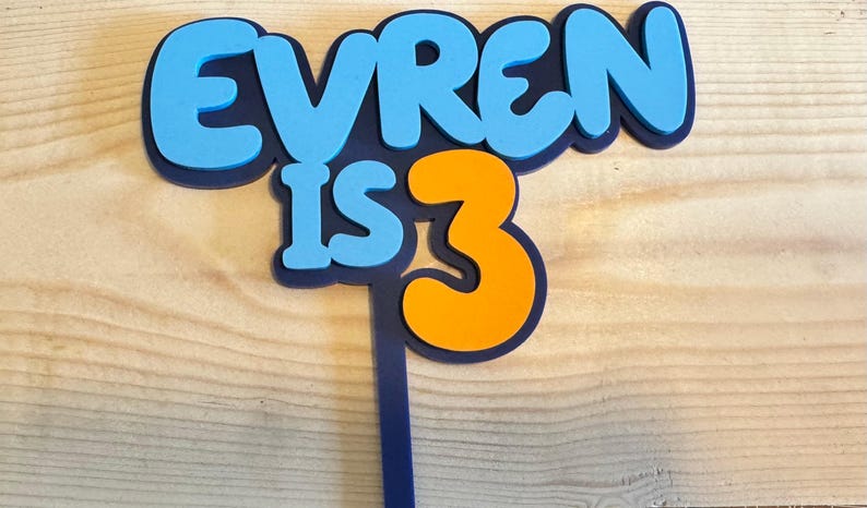 May include: A blue and orange cake topper with the text "EVREN IS 3". The letters are in a bubble font with a dark blue outline. The number 3 is orange. The topper is on a wooden surface.