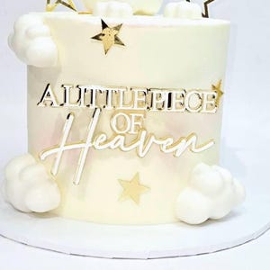 Pode incluir: Um bolo cilíndrico com cobertura branca lisa, decorado com nuvens de pasta americana branca e estrelas douradas. A frente apresenta um topo de bolo em acrílico dourado com o texto A LITTLE PIECE OF HEAVEN em fontes de bloco e cursiva.