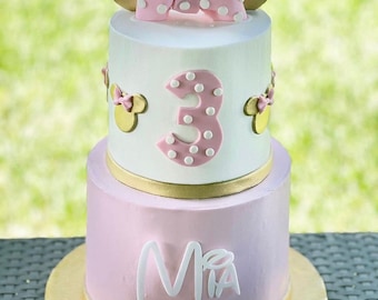 May include: Two-tiered pink and white birthday cake decorated with a pink bow, gold ears, and white polka dots. The cake is decorated with the number "3" and the name "Mia".