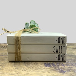 May include: Three white wooden blocks stacked on top of each other. The top block says "HOME", the middle block says "SWEET", and the bottom block says "HOME". A small green plant is sitting on top of the blocks.