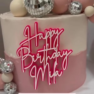 May include: A two-tiered birthday cake with pink and white frosting. The cake is decorated with disco ball and pearl-like ornaments. A pink and white acrylic cake topper reads "Happy Birthday Mifa".