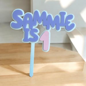 May include: A cake topper in light blue, featuring the text "Sammie is 1" in purple and pink. The letters have a rounded, bubble-like appearance. The topper is designed to be placed on a cake.