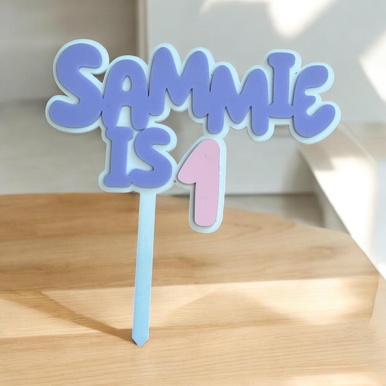 May include: A cake topper in light blue, featuring the text "Sammie is 1" in purple and pink. The letters have a rounded, bubble-like appearance. The topper is designed to be placed on a cake.