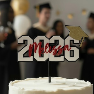 Op de afbeelding: Een caketopper met het jaartal "2026" in zwarte en witte blokletters, met de naam "Melissa" in rood script. Een gouden glitter afstudeerhoed zit boven de cijfers. De topper staat op een zwarte stok, geplaatst op een wit geglazuurde taart.