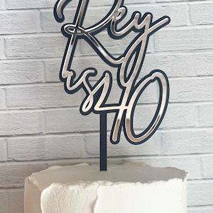 May include: A black and silver cake topper that reads "Rey is 40" in a cursive font. The topper is placed on top of a white frosted cake, with a white brick wall in the background.