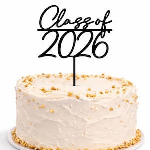 May include: A white iced cake with gold sprinkles and a black cake topper reading "Class of 2026". The cake sits on a white plate, ideal for a graduation party.