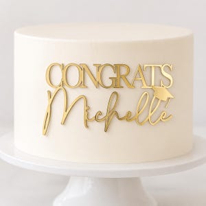 Könnte beinhalten: Ein weißer Kuchen auf einem weißen Kuchenständer. Der Kuchen ist mit goldenen Buchstaben verziert, die "CONGRATS Michelle" und eine Abschlusskappe zeigen. Ein festliches Dessert für eine Abschlussfeier.
