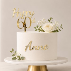 May include: A white cylindrical cake featuring a gold-colored acrylic cake topper that reads Happy 60th and a gold-colored acrylic name plaque that reads Anne. The cake is decorated with a white rose, small white flowers, and green leaves.