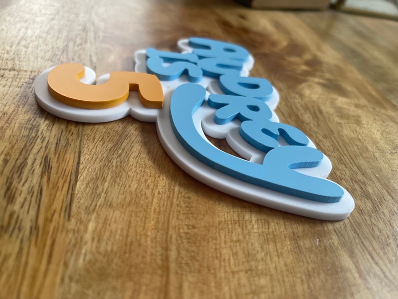 May include: A layered sign with the words "5 more" in a playful font. The sign features a white base, light blue lettering, and an orange number 5. The sign is on a wooden surface.