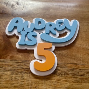 May include: A layered cake topper with the words "Audrey is 5" in blue and orange. The text is layered on a white base, creating a 3D effect. The number 5 is in orange. The topper is designed for a birthday celebration.